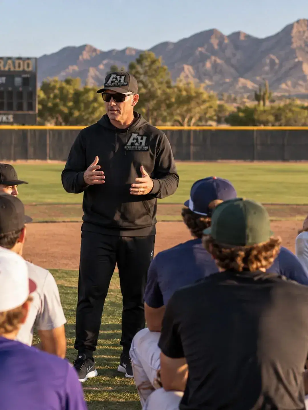 BK talking to a circle of baseball players on the field