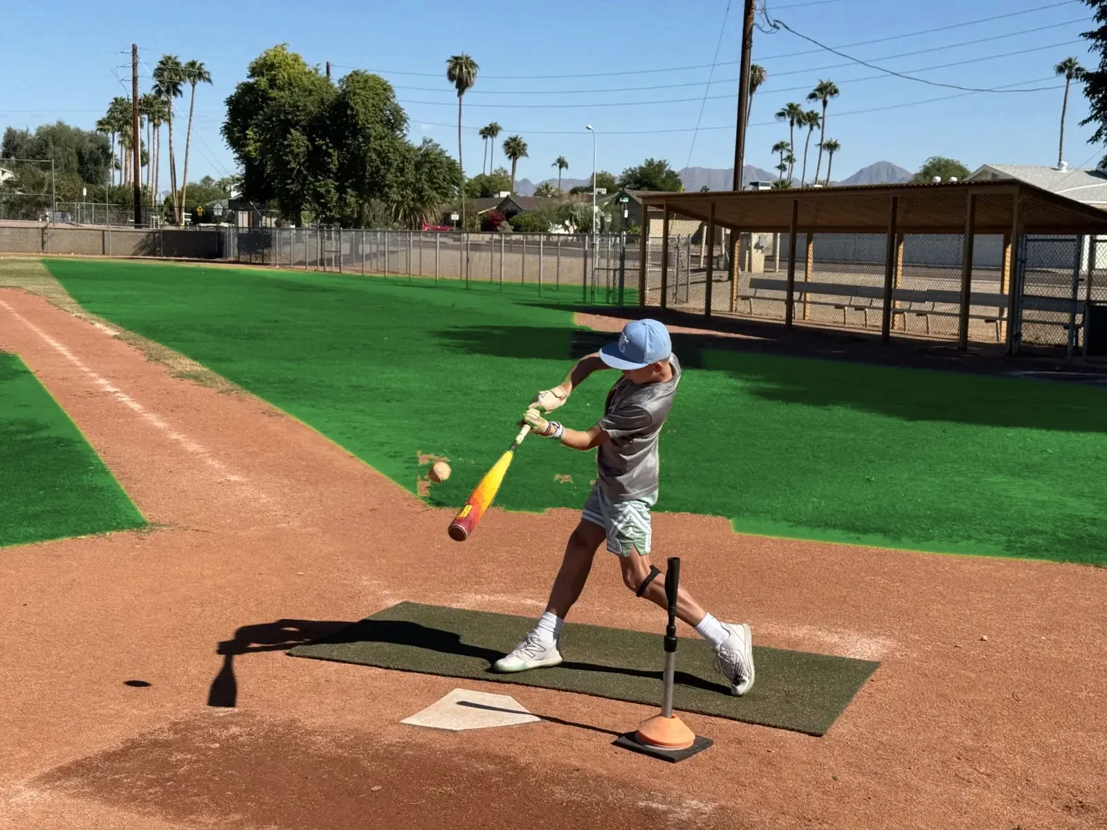 Young hitter driving a ball off the tee on a Scottsdale field with palm trees and desert mountains under a blue sky.